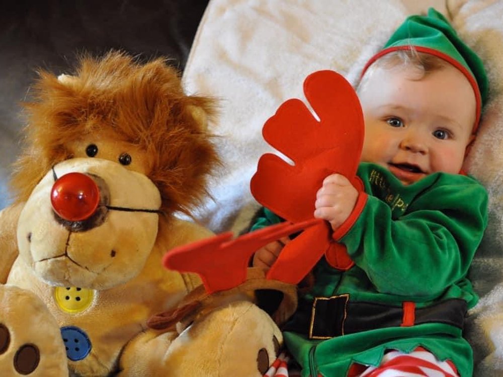A baby with mitochondrial disease wearing a cute green elf outfit and holding a reindeer soft toy