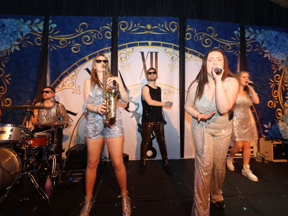 A band wearing sparkly clothing on stage at the Lily ball, one is singing into a microphone, others playing drums and guitar