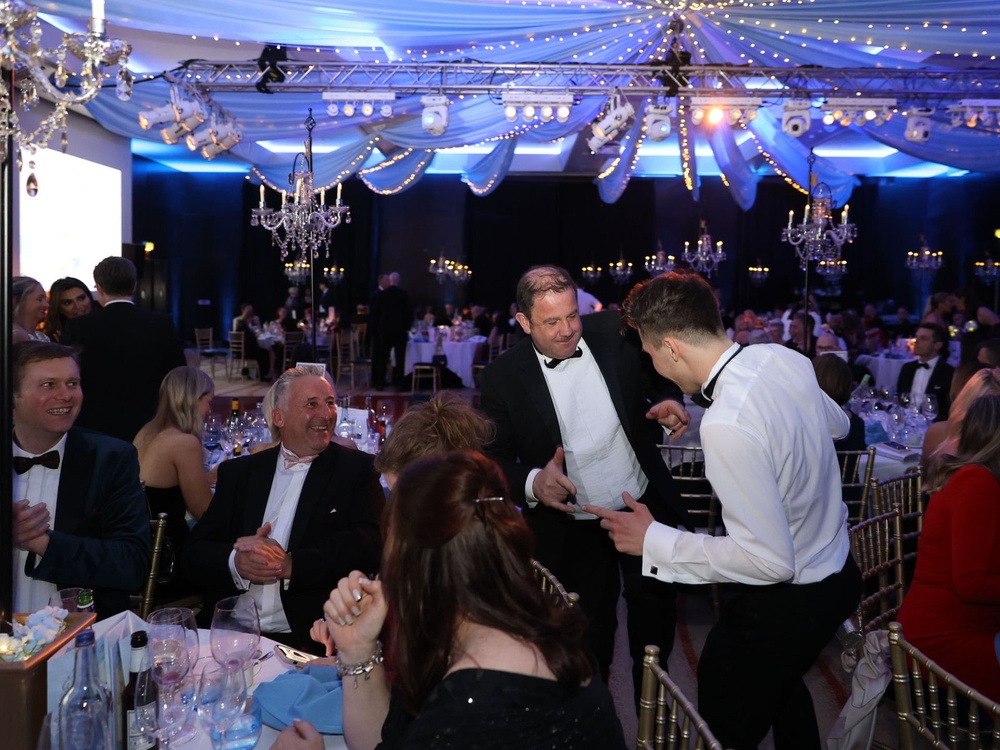 Guests sitting at their tables and enjoying themselves with two men in black tuxedos standing up laughing