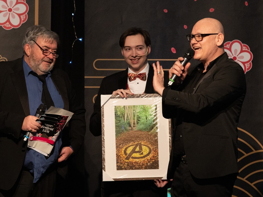 Three men stand on stage with a picture, one speaking into a microphone