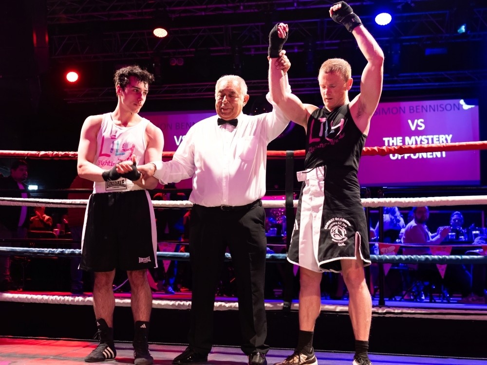 A boxer holds his hands up victorious next to the ref and his opponent