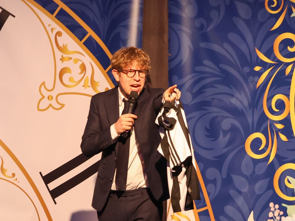 Comic Josh Widdicombe standing on stage with a microphone and pointing