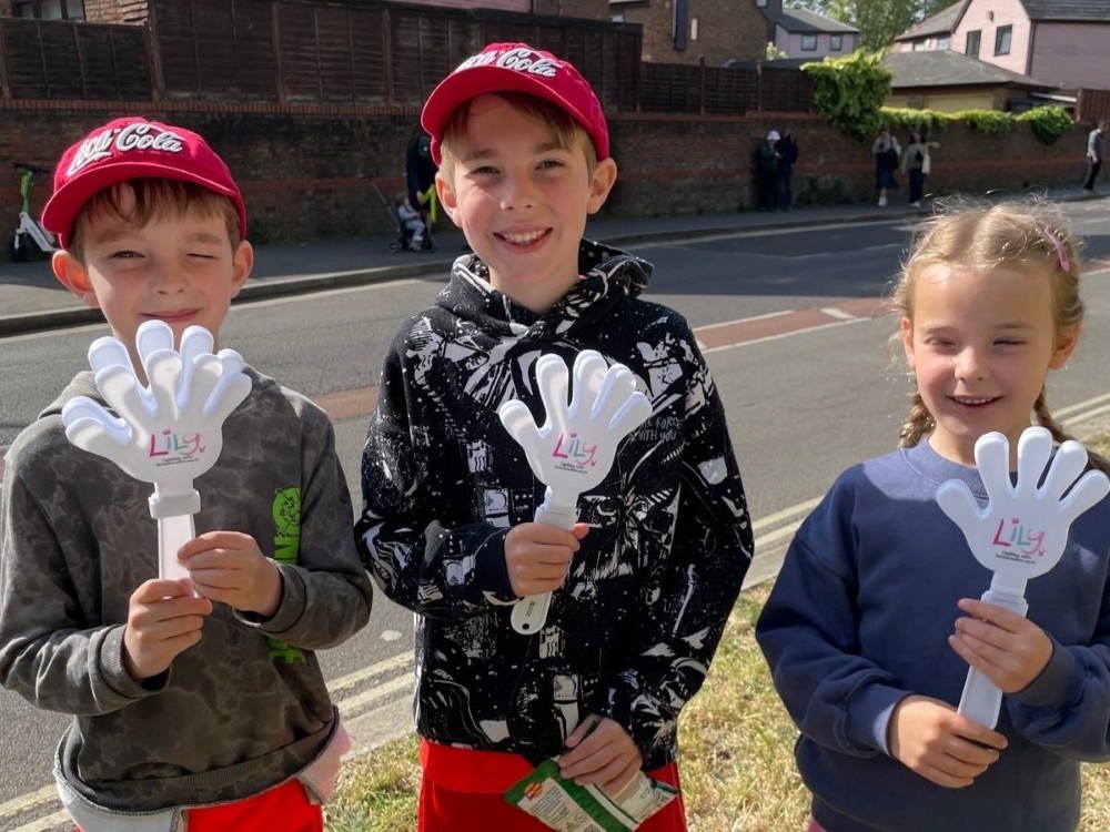 Three young children wearing Lily tops and holding clappers by the side of the road