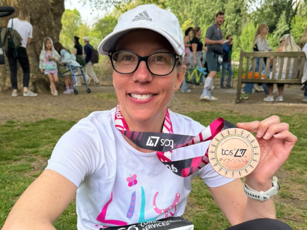 A lady in a Lily top and baseball cap holding her London marathon medal up proudly