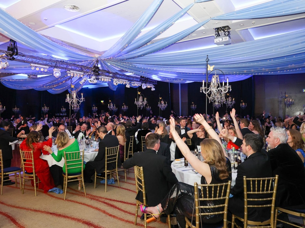 Guests seated at tables at the Lily ball holding their arms up in the air and cheering