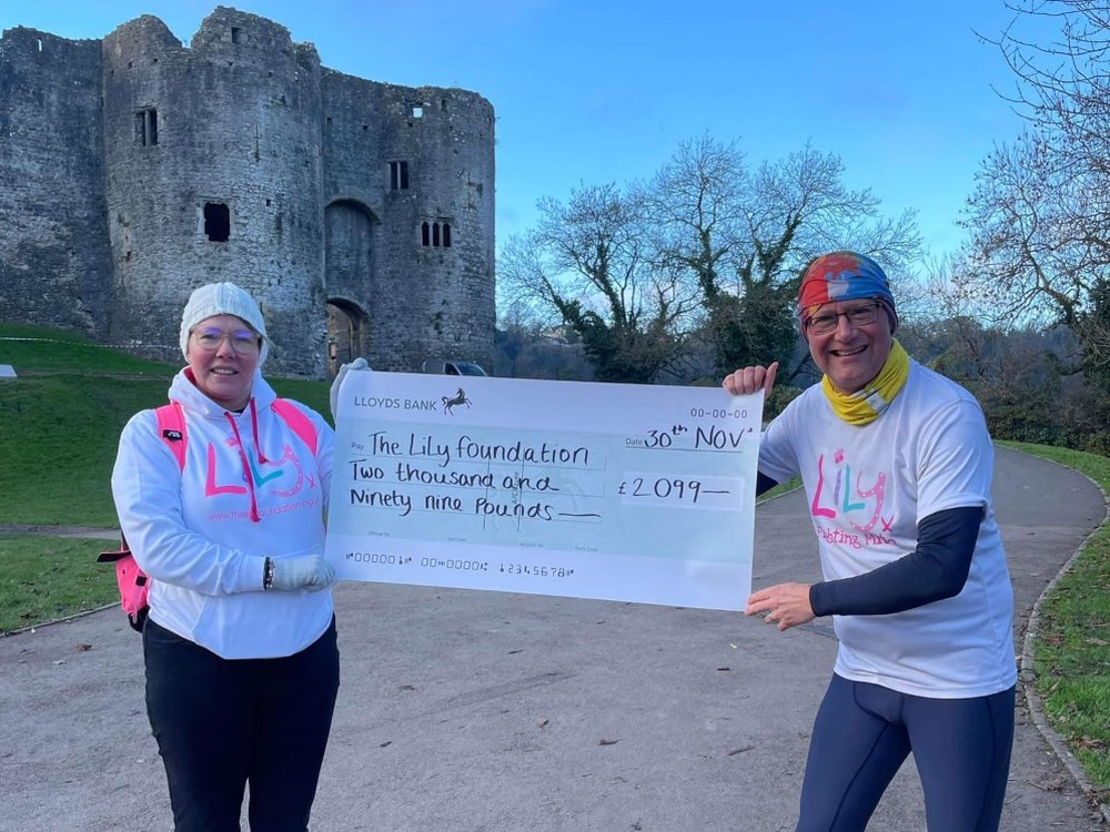 A man and woman in Lily tops holding a large cheque and standing in front of a castle
