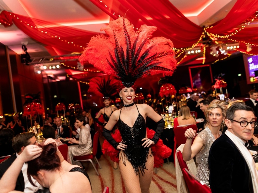 A showgirl dressed in black with red feathers strutting past tables at the Lily ball