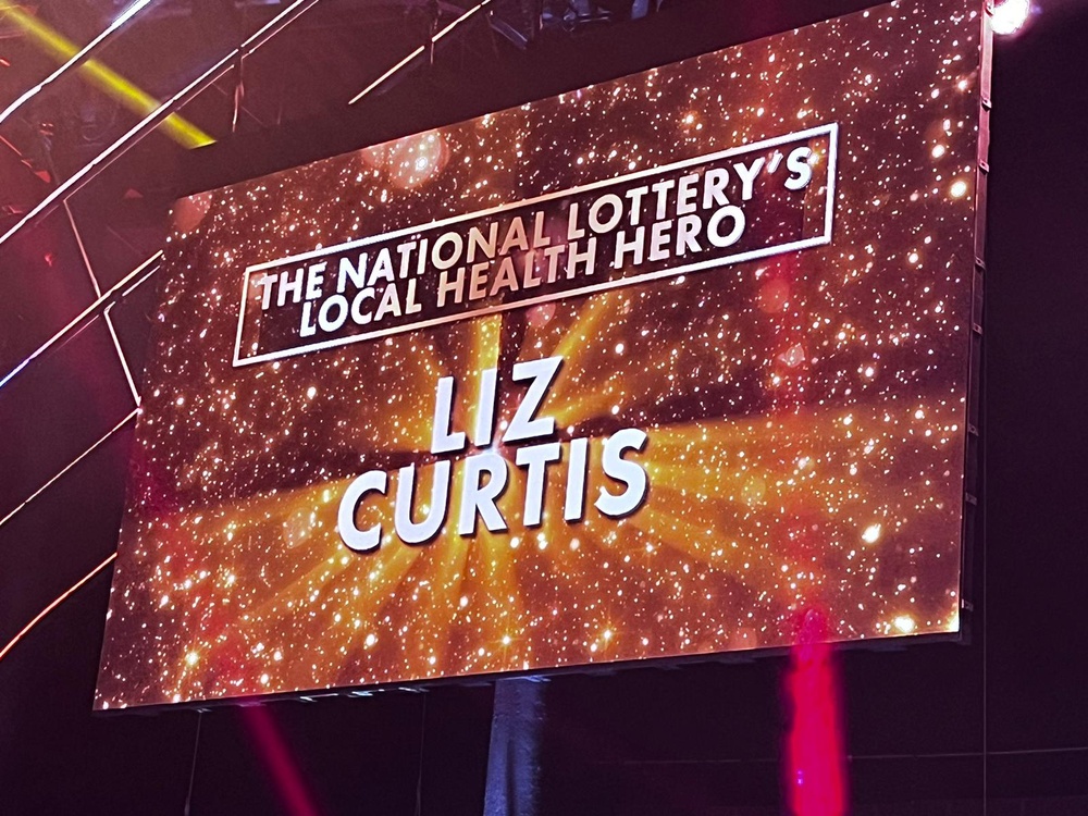 A giant TV screen showing the name of Liz Curtis as winner of the National Lottery Local Health Hero award