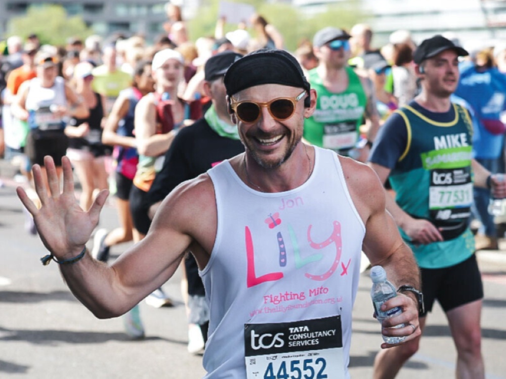 A man in a Lily vest and sunglasses running the London marathon waves at the camera