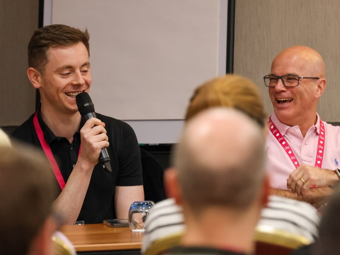 Two men are seated behind a desk, one speaking into a microphone, to a group of adults in front of them