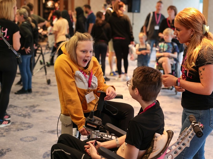 Two children, a girl and a boy, in wheelchairs are laughing and exclaiming together. Both are wearing Lily lanyards.