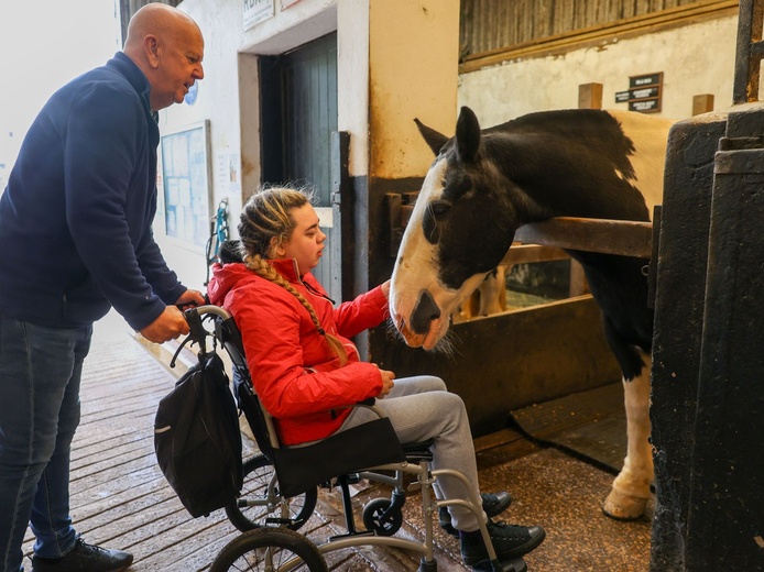 A young lady in a wheelchair strokes a horse's face in a stable