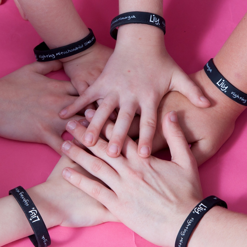 A group of people's hands stacked on top of each other, all wearing a black wristband featuring the Lily Foundation logo.