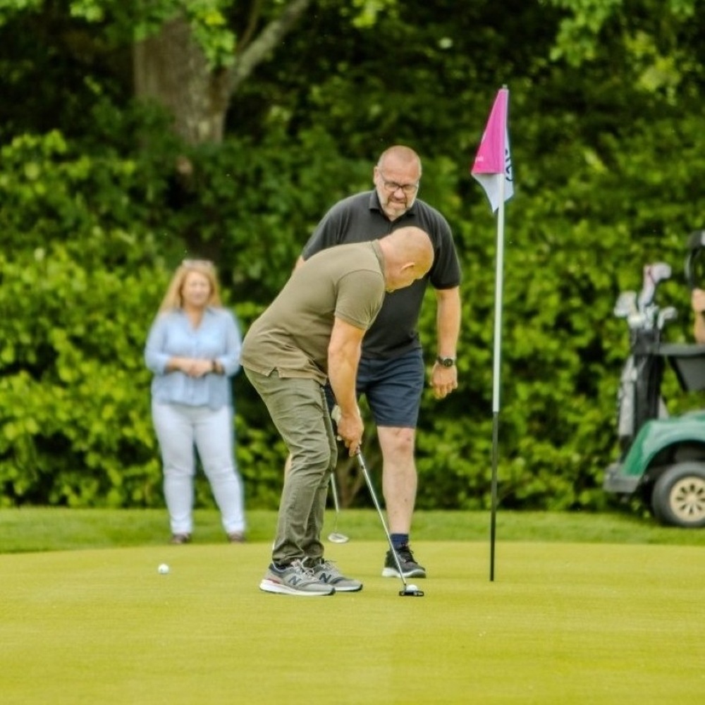 A man on the green of a golf course taking a shot, with another man standing next to the hole