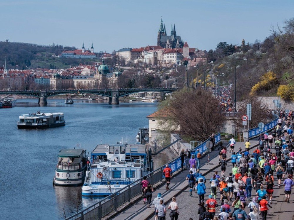 The image shows a beautiful Prague scene with crowds of runners on a path along the edge of the River.
