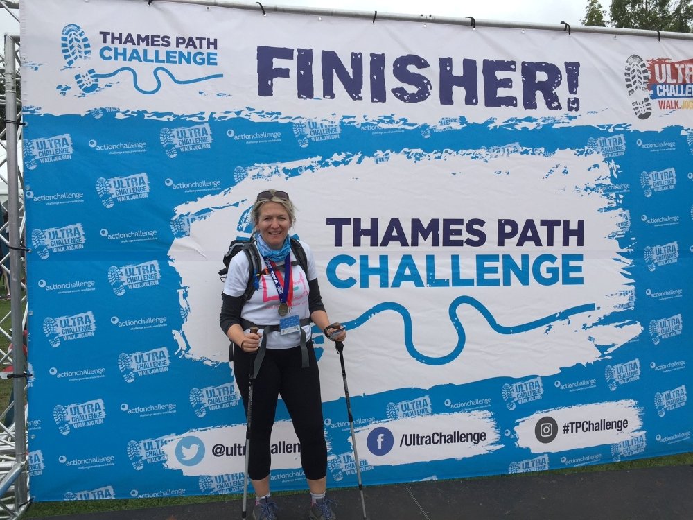 A lady in a Lily Foundation top holding walking poles and standing in front of the Thames Path challenge finishers' banner