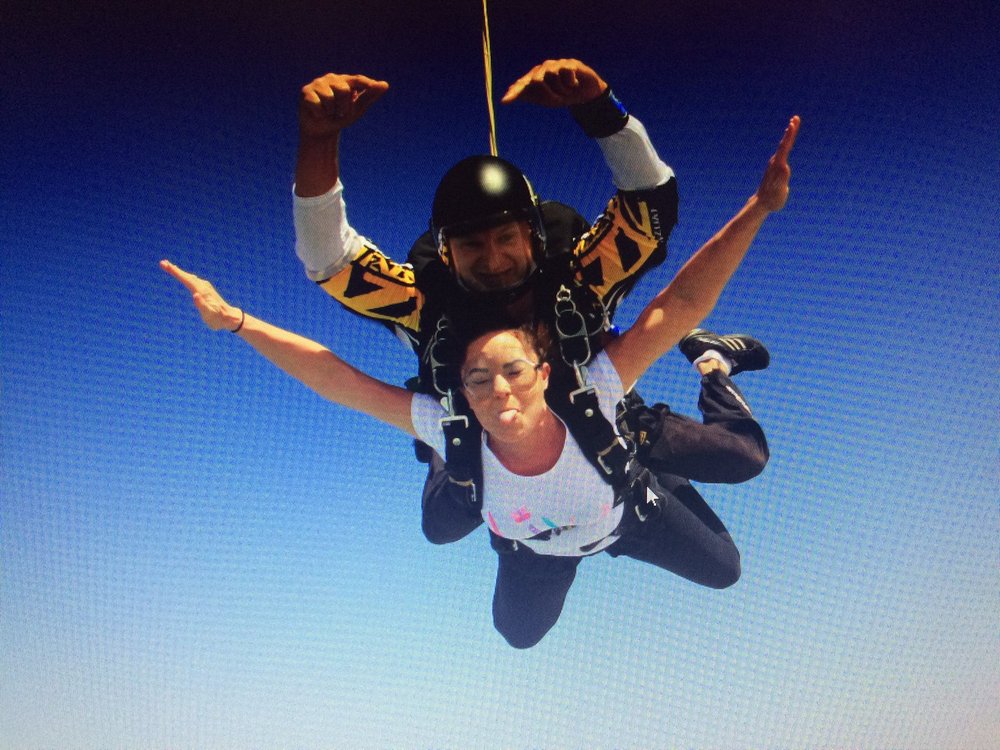 Two people on a parachute jump falling through the air with blue sky behind, one is wearing a Lily Foundation top and raising her arms