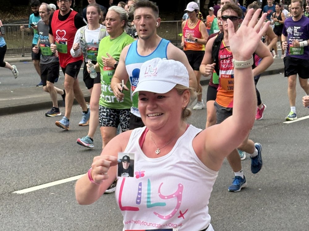 A woman running the Great North Run, wearing a Lily vest, waving and holding a photo of her loved one