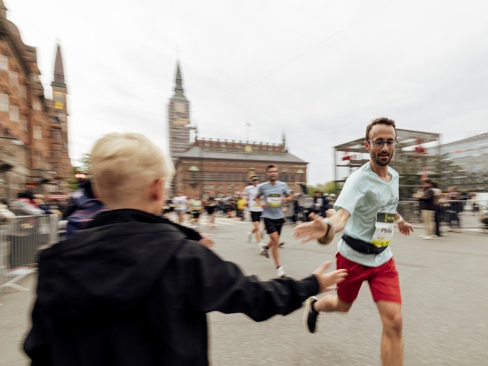 The image shows a male runner in t-shirt and shorts being supported by a young spectator as he runs the Copenhagen half marathon