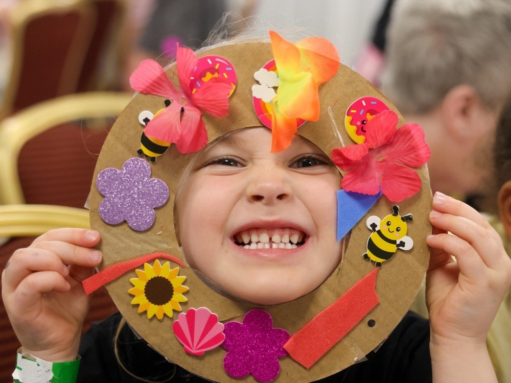 Lily Family Support Weekend 2027 – Save the Date - A smiling boy peers through a face mask decorated with colourful flowers