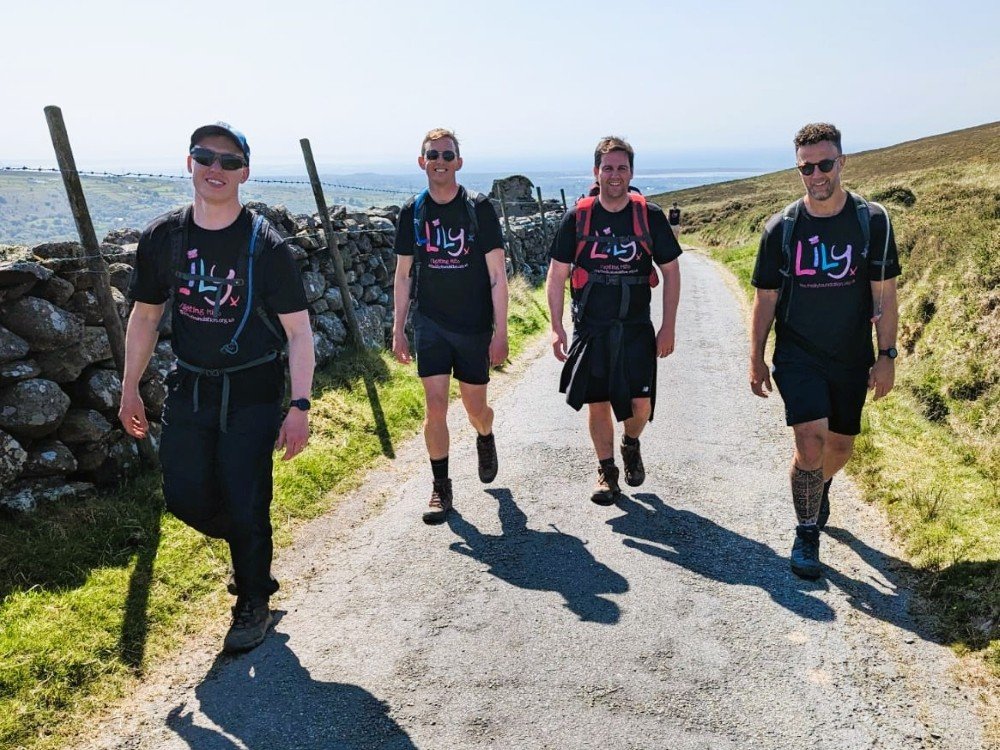 Four men in Lily Foundation tops walking in the countryside with hills and mountains in the background.