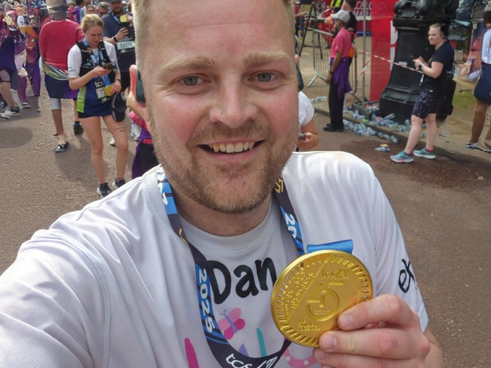 Man with light brown short hair and a short beard wearing a white Lily Foundation t-shirt. He is smiling and holding his London Marathon finishers medal in a selfie photograph.