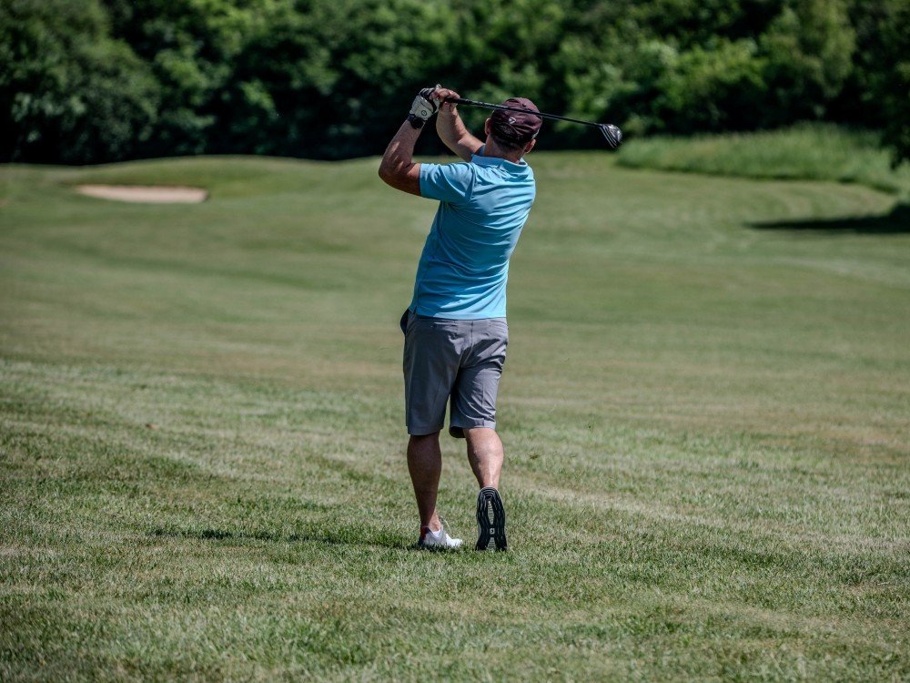Lily South West Golf Day 2027 – Save the Date - A man in a blue t-shirt and dark shorts swings his golf club on the green at the Lily South West Golf Day