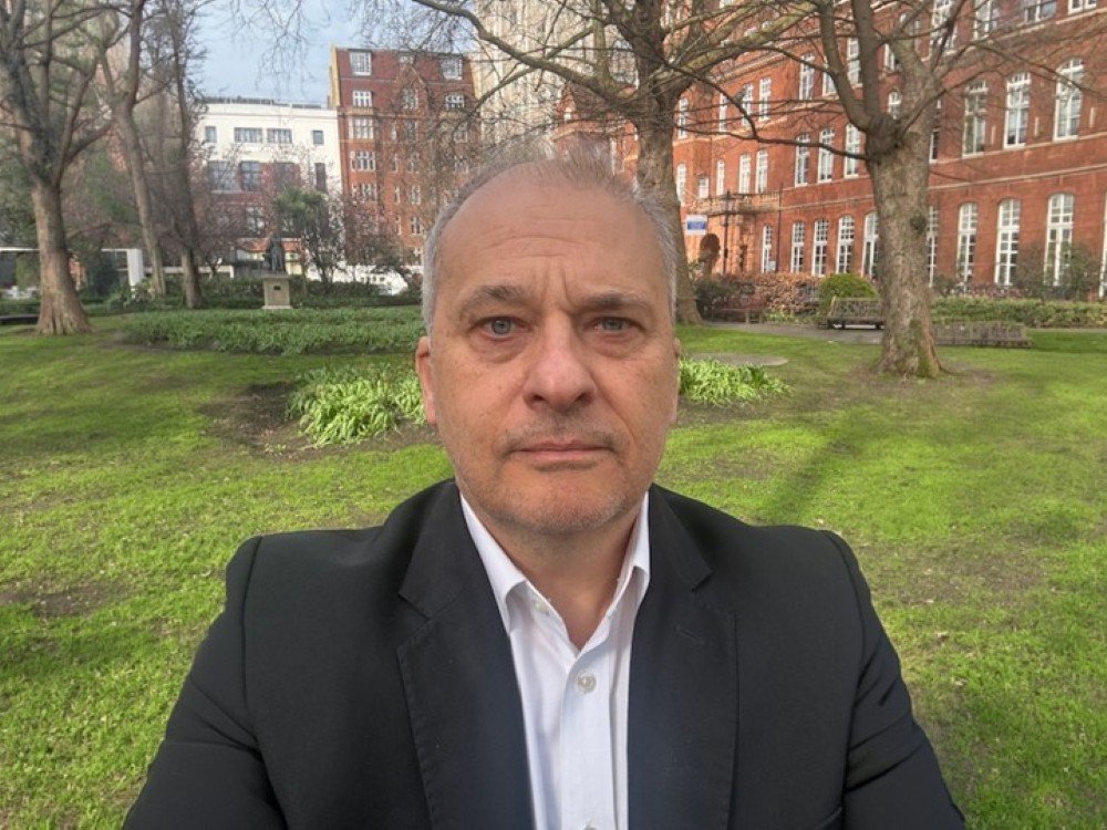 Close-up of Professor Mike Hanna, director of the UK Mitochondrial Disease Research Institute, in a park with buildings behind