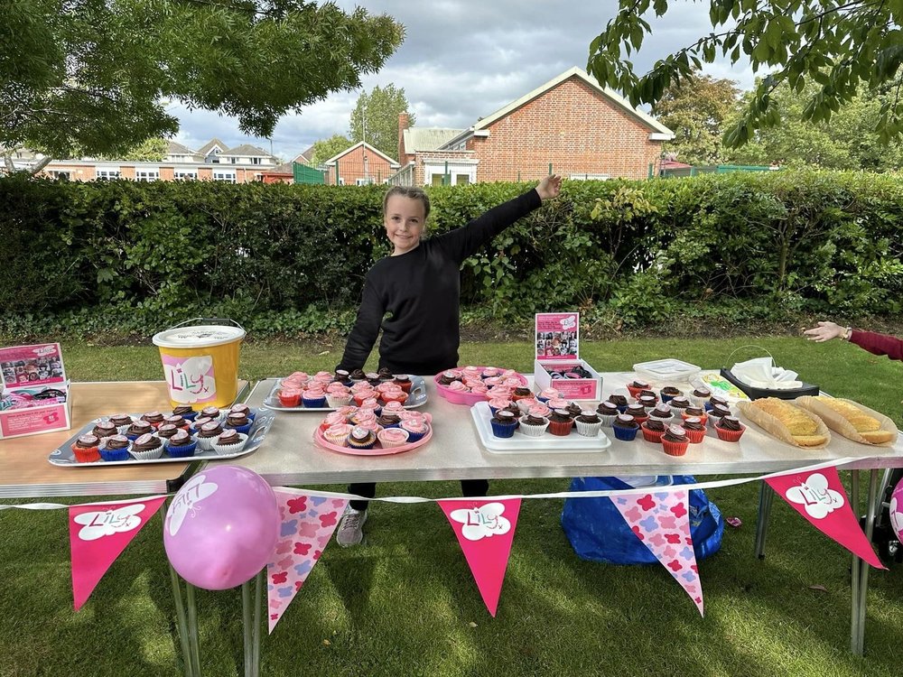 a young girl with her thumbs up, manning a Lily fundraising cake stand, fully of delicious cakes