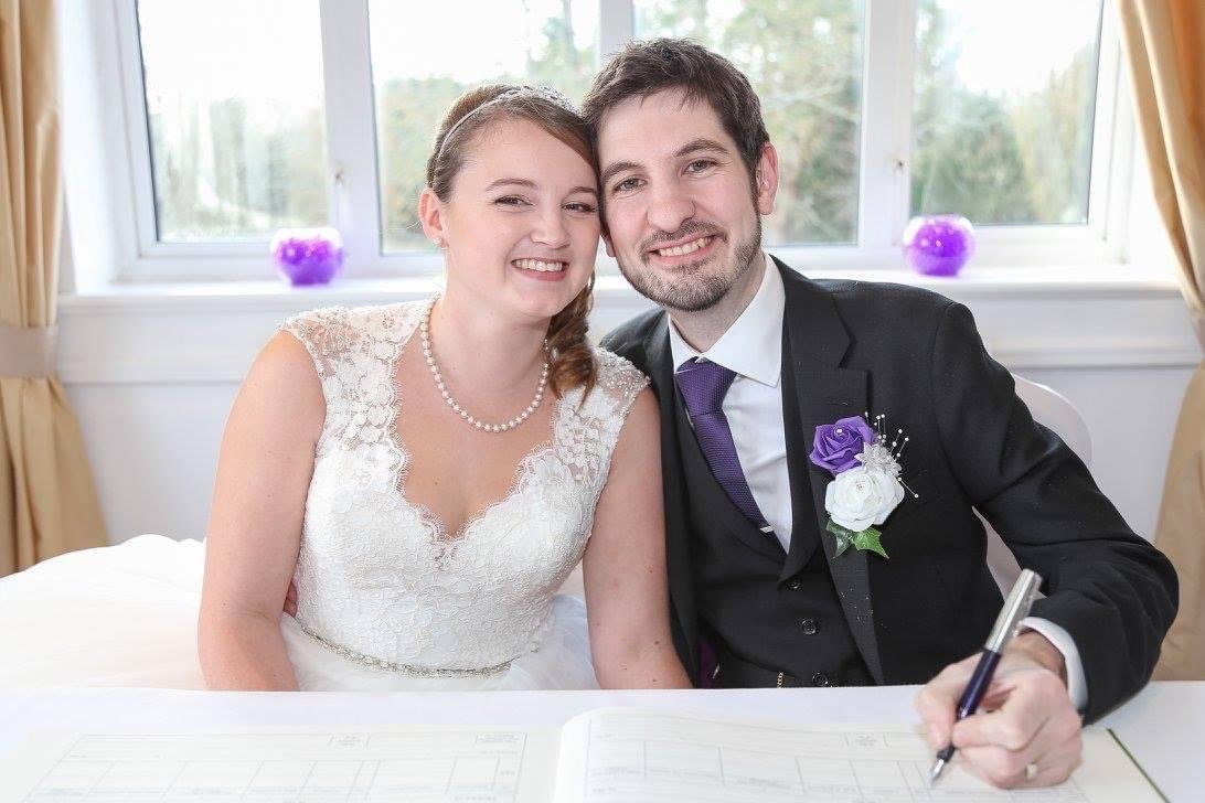 Josey in her wedding dress and Chris in suit and tie signing their wedding certificate and beaming at the camera