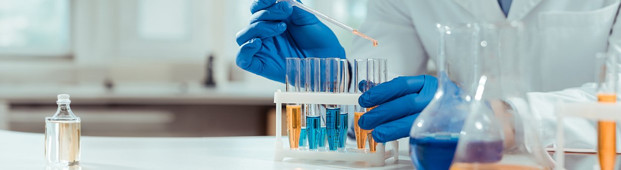 Lab technician wearing blue gloves, using a pipette to  put blue or yellow liquid into  a rack of test tubes