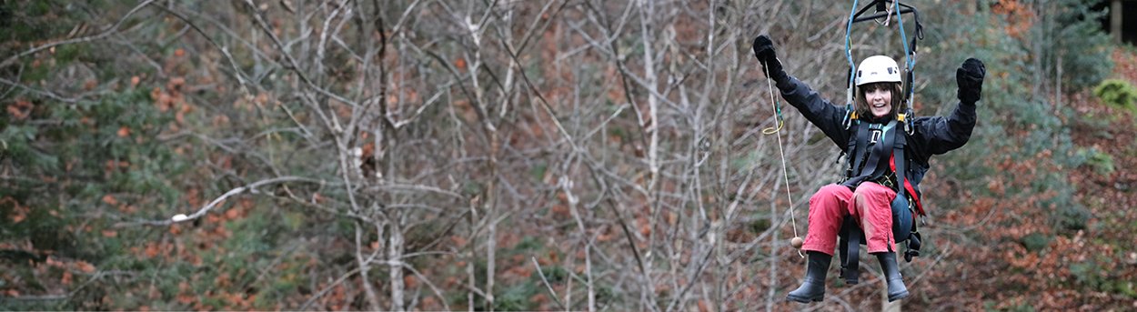 A lady wearing a crash helmet zipwiring through trees grinning with her hands in the air