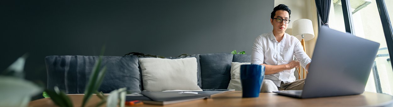 Man sitting on the end of a grey sofa, engaging with someone via his open laptop