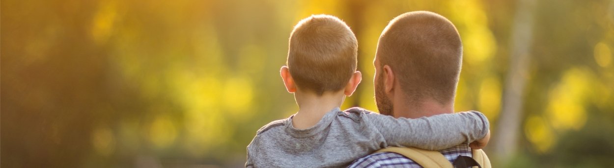 A man holding his son in his arms stands with his back to the camera gazing at trees