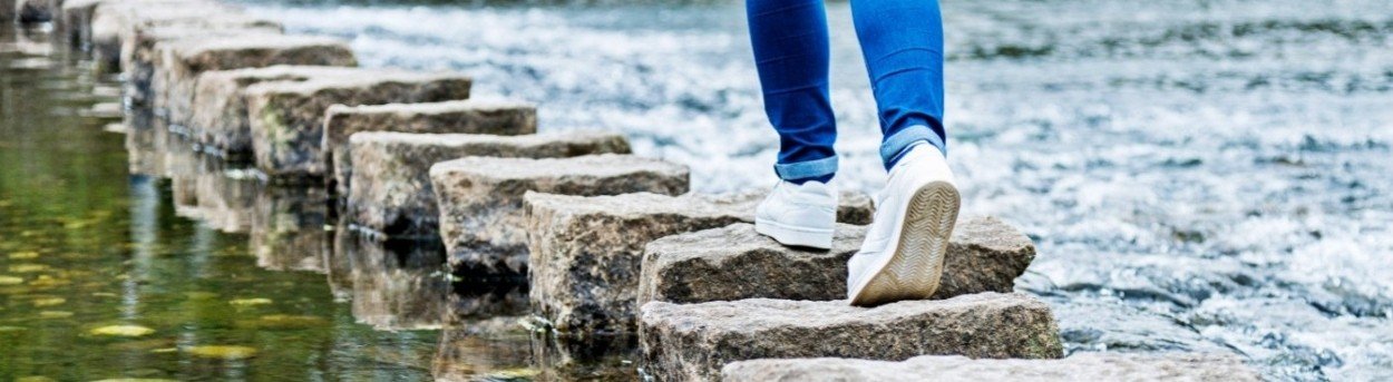 Close up of someone wearing jeans and trainers walking across stepping stones over a river