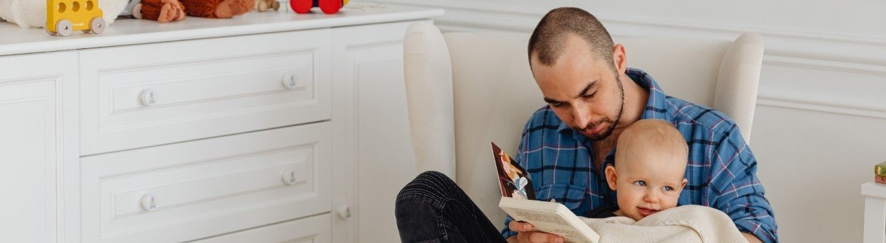 A man seated on a white chair reading to a young baby in his lap under a cream blanket