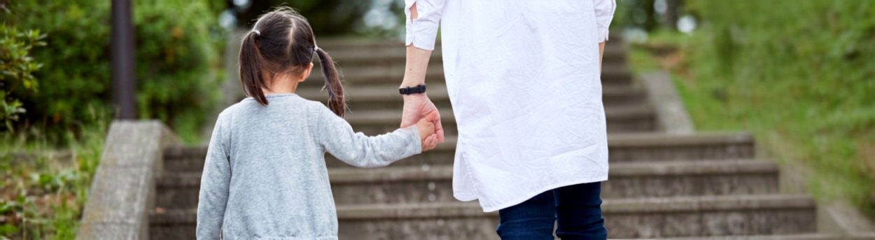 An adult in a white coat holding the hand of a small child as they climb some steps outdoors