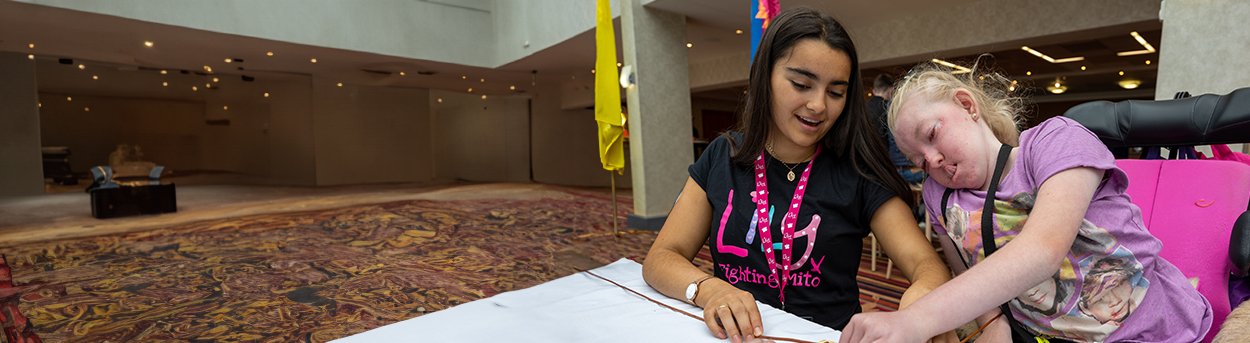 Two girls, one in a Lily t-shirt and lanyard, the other in a wheelchair, working at a table