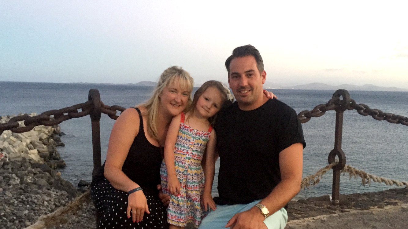 A man and women sit with a little girl in the middle of them in front of the ocean smiling at the camera