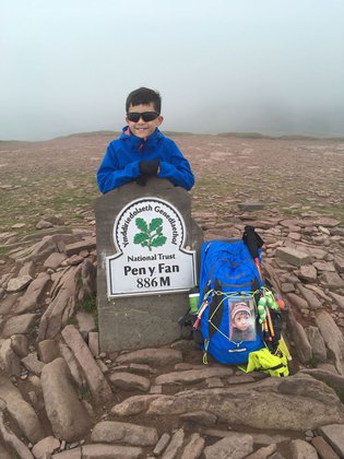 A  boy stands behind a sign that says Pen y Fan next to the sign is a blue ruck sack with a picture of a little boy on it