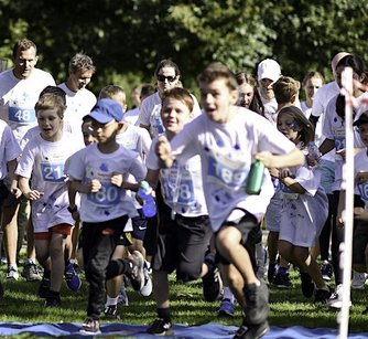 A group of children wearing running tops with numbers eagerly setting off from the start line of a race, with adults behind