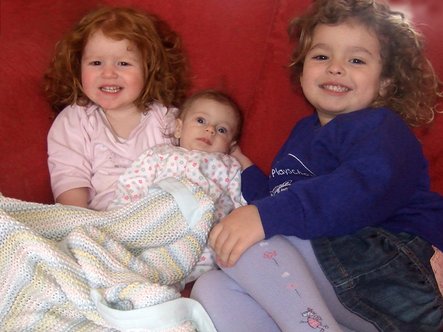 Baby Lily having a cuddle on a red sofa with her sisters Rosie and Katie