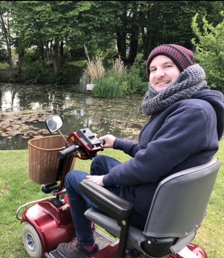 Chris in his wheelchair in front of a pond turning to the camera