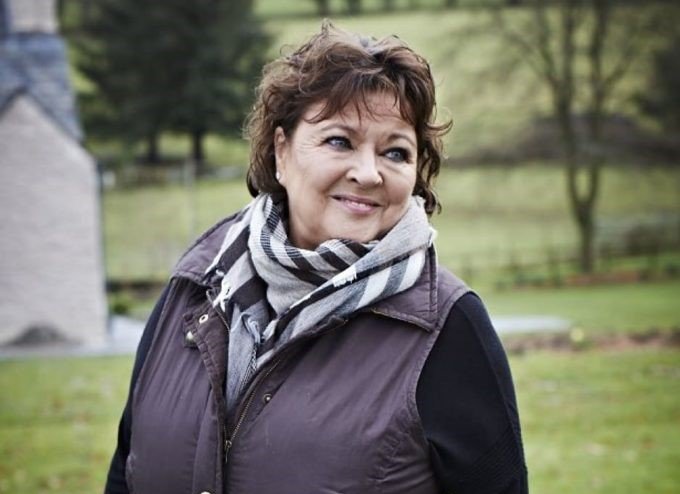 A lady with short dark hair standing in a field wearing a gilet and scarf, laughing and looking just off camera
