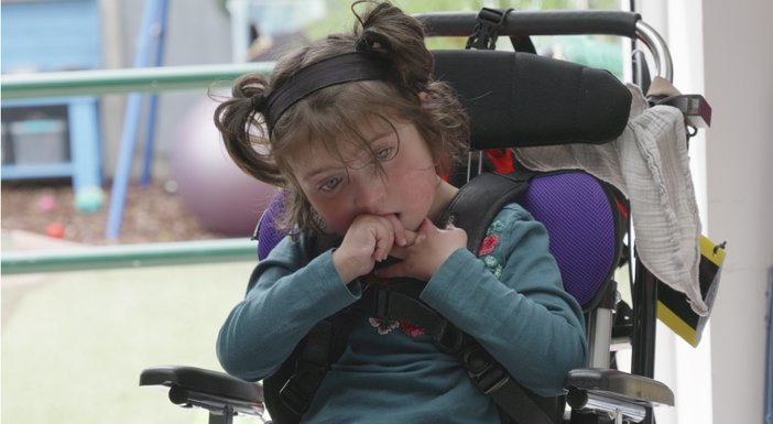 A young mitochondrial disease patient in a wheelchair holding her hands to her mouth