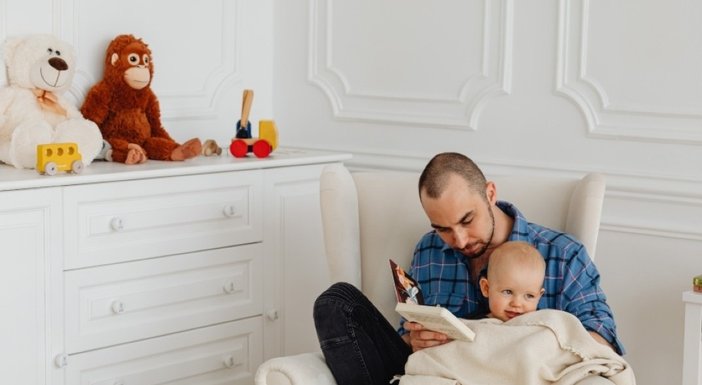 A man seated on a white chair reading to a young baby in his lap under a cream blanket