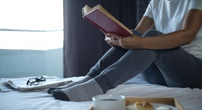A person sitting in their socks on a bed reading a book