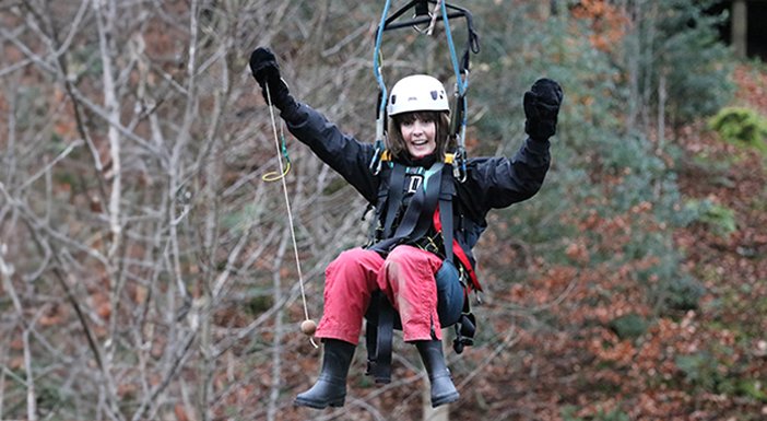 A lady wearing a crash helmet zipwiring through trees grinning with her hands in the air