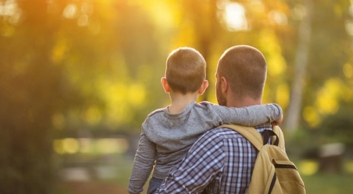 A man holding his son in his arms stands with his back to the camera gazing at trees