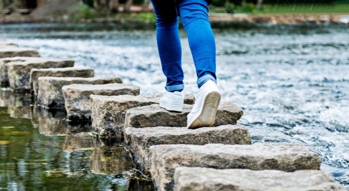 Close up of someone wearing jeans and trainers walking across stepping stones over a river